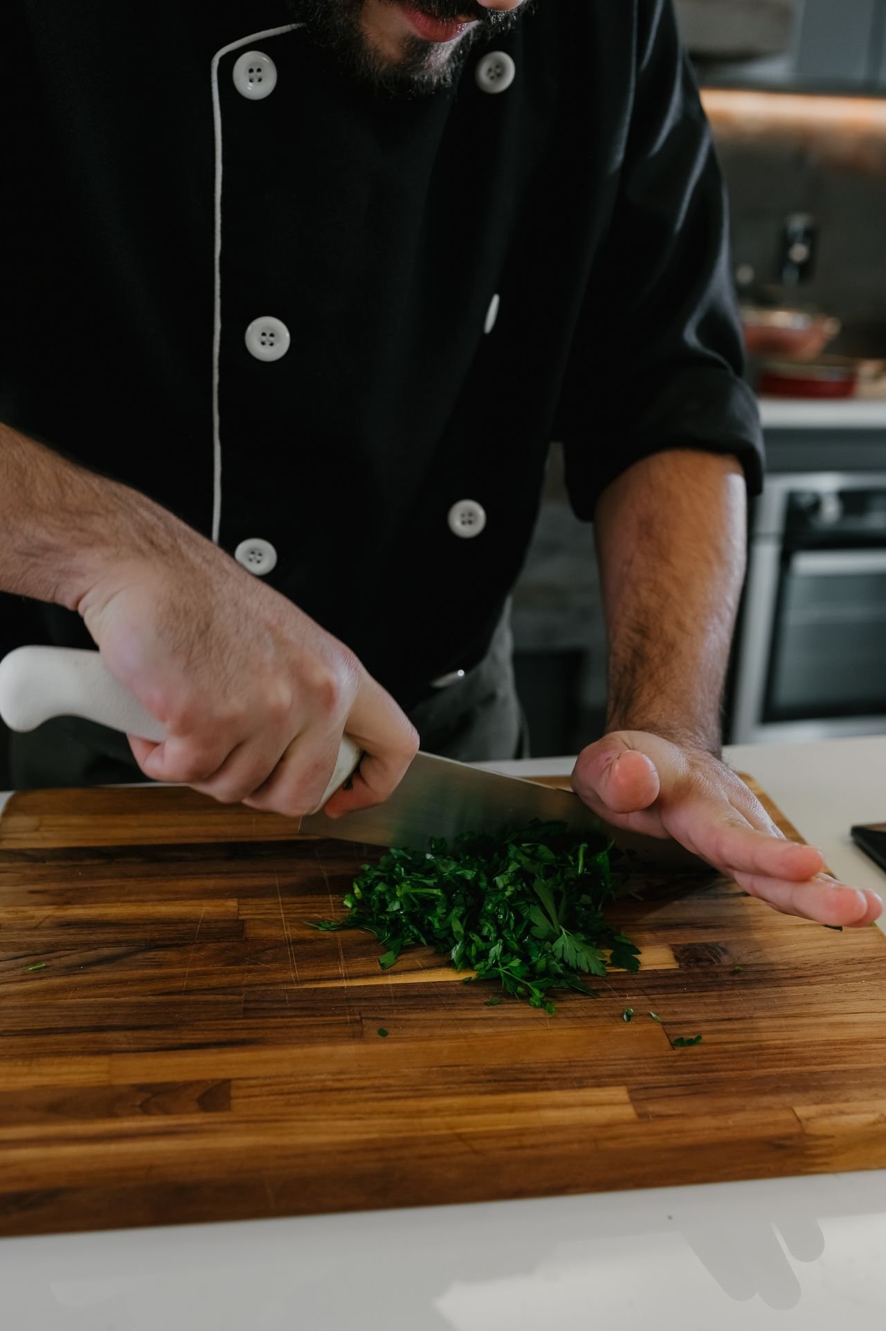 Chef con uniforme negro cortando hierbas frescas con un cuchillo tradicional sobre tabla de madera